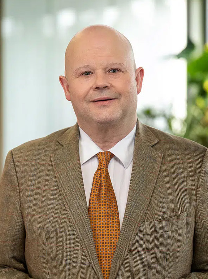 Portraitfoto eines Mannes mit Glatze, bräunlichem Anzug und oranger Krawatte