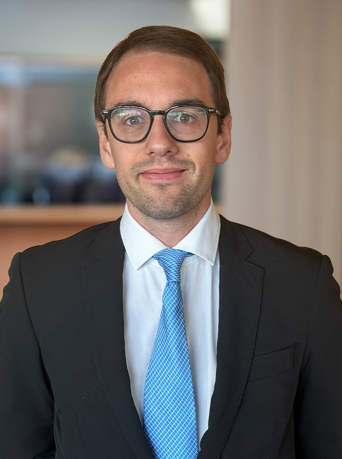 Porträt eines Mannes mit kurzen braunen Haaren und schwarzer Brille, der einen dunklen Anzug, ein weißes Hemd und eine blaue Krawatte trägt.