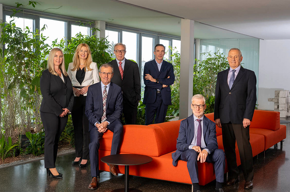 Eine 7-köpfige Gruppe Frauen und Männer steht und sitzt bei einem orangen Sofa