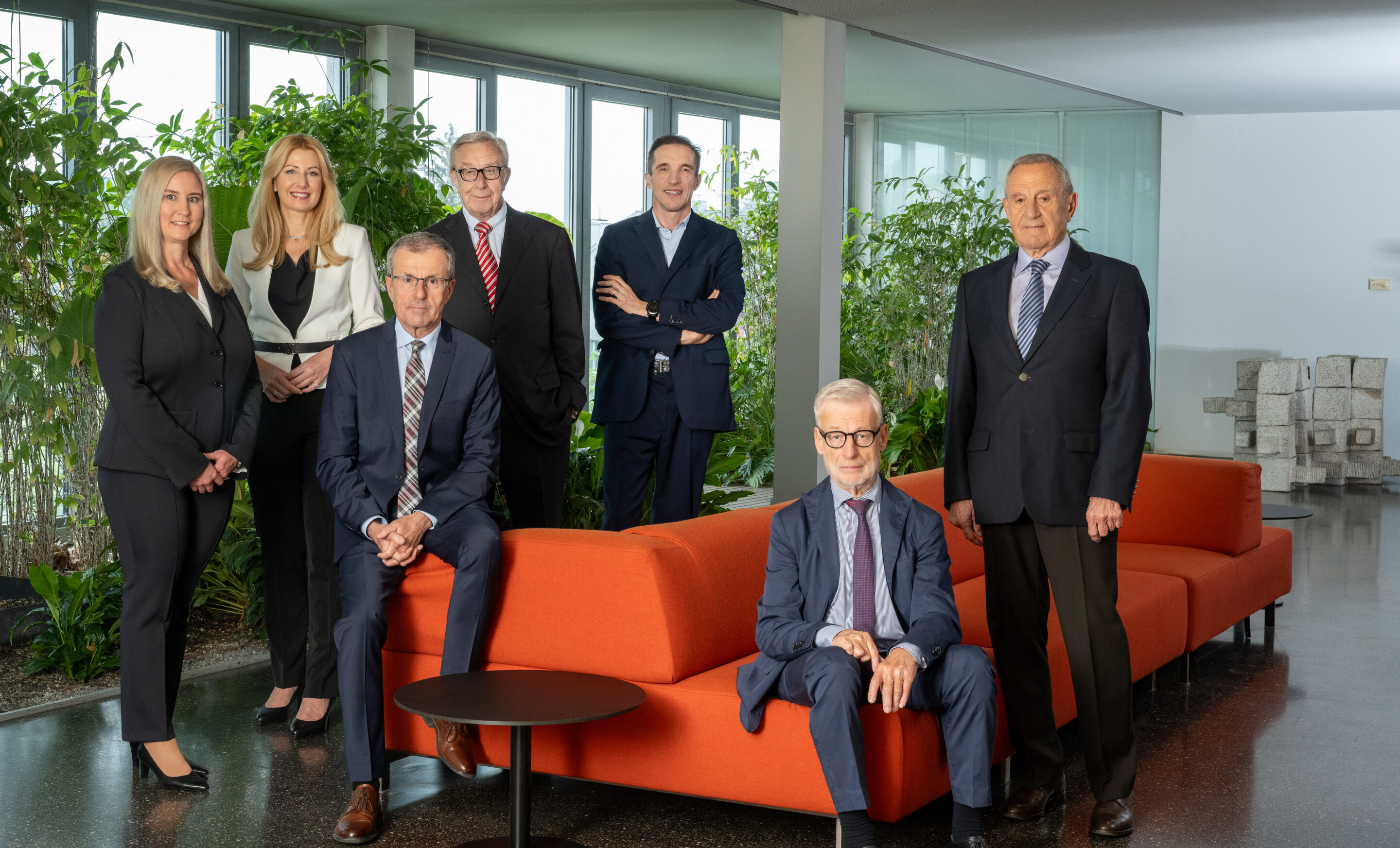 Eine 7-köpfige Gruppe Frauen und Männer steht und sitzt bei einem orangen Sofa
